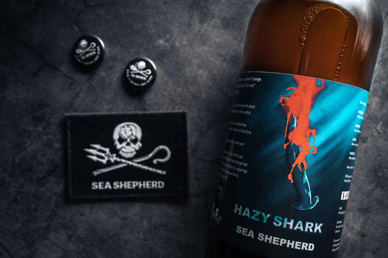 Gros plan sur l'étiquette d'une bouteille de bière Sea Shepherd Hazy Shark IPA, patch avec logo et deux pins Sea Shepherd Don't drink and dive
