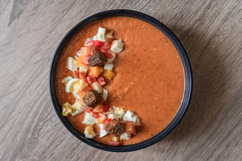 Bol de gaspacho au pain avec des croutons de pain aux drêches, de l'oeuf dur et du fromage de chèvre.